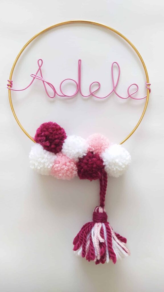 décoration murale avec un cercle doré avec prénom en fil aluminium rose et pompons en laine couleurs bordeaux, rose et blanc