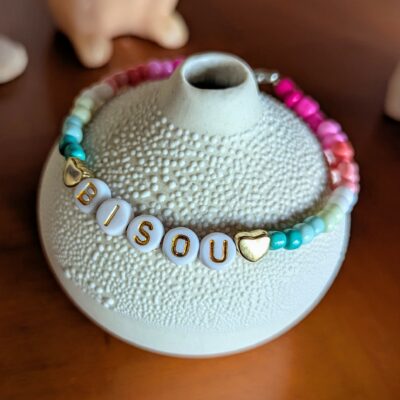 Bracelet en perles de rocailles en dégradé allant du bleu au rose avec 2 perles coeur, mot "bisou" écrit en perles plastique dorées. Avec fermoir argenté et chainette.