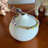 L’image montre un petit vase rond en céramique blanche, à la surface texturée en relief, posé sur une table en bois. Autour du col du vase est délicatement posé un bracelet coloré composé de perles rondes. Au centre du bracelet, on distingue des perles lettres blanches avec des lettres noires formant le mot "MORUE", encadré de deux perles avec des cœurs colorés (rose à gauche, orange à droite). Les autres perles du bracelet sont de différentes couleurs vives : jaune, violet, vert, rose, blanc… En arrière-plan, on aperçoit une figurine animale floue (probablement un éléphant), une décoration en forme de gland, et d'autres objets décoratifs, donnant une ambiance chaleureuse et artisanale à la scène.