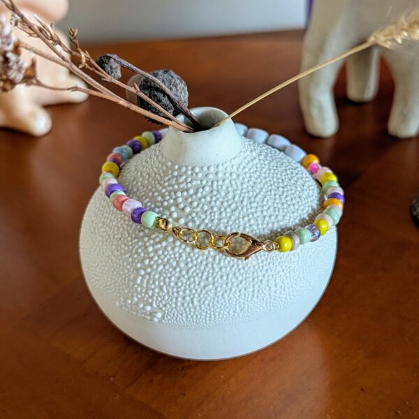 L’image montre un petit vase rond en céramique blanche, à la surface texturée en relief, posé sur une table en bois. Autour du col du vase est délicatement posé un bracelet coloré composé de perles rondes. Au centre du bracelet, on distingue des perles lettres blanches avec des lettres noires formant le mot "MORUE", encadré de deux perles avec des cœurs colorés (rose à gauche, orange à droite). Les autres perles du bracelet sont de différentes couleurs vives : jaune, violet, vert, rose, blanc… En arrière-plan, on aperçoit une figurine animale floue (probablement un éléphant), une décoration en forme de gland, et d'autres objets décoratifs, donnant une ambiance chaleureuse et artisanale à la scène.