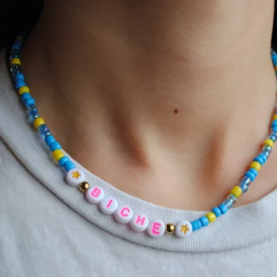 Joli collier fantaisie en perles de rocaille bleu et jaune, avec petites perles étoiles jaunes, et mot "biche" écrit en lettre roses fluos.