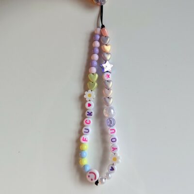 On voit une dragonne de téléphone faite main, suspendue par un cordon noir tenu entre deux doigts. Elle est composée de deux brins de perles aux couleurs pastel, avec des éléments décoratifs ludiques et féminins. Le brin de gauche est orné de perles rondes dans des tons doux (bleu, jaune, rose, blanc), de perles en forme de marguerites blanches, et de lettres qui forment le mot "FUCK", avec un petit cœur rouge à la fin. Tout en bas, une perle blanche affiche un smiley rose légèrement sarcastique. Le brin de droite est plus scintillant, avec des perles en forme de cœurs dorés et nacrés, une étoile irisée, une petite perle rose avec une étoile rouge, ainsi que les lettres formant "YOU", terminées également par un petit cœur rouge. Une grande perle nacrée et d’autres perles transparentes ajoutent de la brillance. L’ensemble repose sur un fond blanc épuré, mettant en valeur les couleurs pastel et les formes mignonnes de la dragonne. Le contraste entre le message audacieux et le style visuel doux crée un effet à la fois drôle et provocateur.