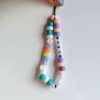 On voit une dragonne de téléphone fait main composé de perles colorées en plastique. Il est formé de deux rangées principales de perles enfilées sur un cordon noir. La rangée de gauche contient des perles blanches, vert turquoise, orange et violettes, ainsi que deux perles en forme de marguerite. Elle porte également des lettres colorées formant le mot "MOULE". La rangée de droite est composée de perles dans des tons pastel (violet, pêche, blanc nacré) et contient des perles blanches avec des lettres noires formant le mot "ALPHA". L'ensemble est tenu en haut par une main avec un ongle verni pailleté.