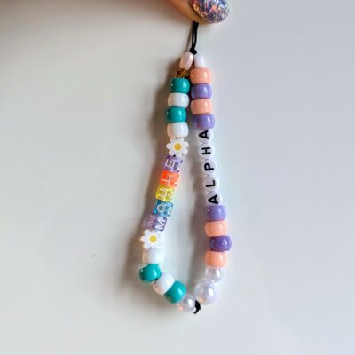 On voit une dragonne de téléphone fait main composé de perles colorées en plastique. Il est formé de deux rangées principales de perles enfilées sur un cordon noir. La rangée de gauche contient des perles blanches, vert turquoise, orange et violettes, ainsi que deux perles en forme de marguerite. Elle porte également des lettres colorées formant le mot "MOULE". La rangée de droite est composée de perles dans des tons pastel (violet, pêche, blanc nacré) et contient des perles blanches avec des lettres noires formant le mot "ALPHA". L'ensemble est tenu en haut par une main avec un ongle verni pailleté.