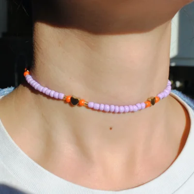 Une personne porte un collier ras-de-cou composé de perles de rocaille violettes, entrecoupées de perles orange vif et de petites breloques en forme de cœur doré. Le collier est ajusté autour du cou, capturant la lumière du jour, ce qui fait ressortir l’éclat des perles et le contraste des couleurs. La personne porte un t-shirt blanc, mettant en valeur les teintes pop et le style délicat du bijou.