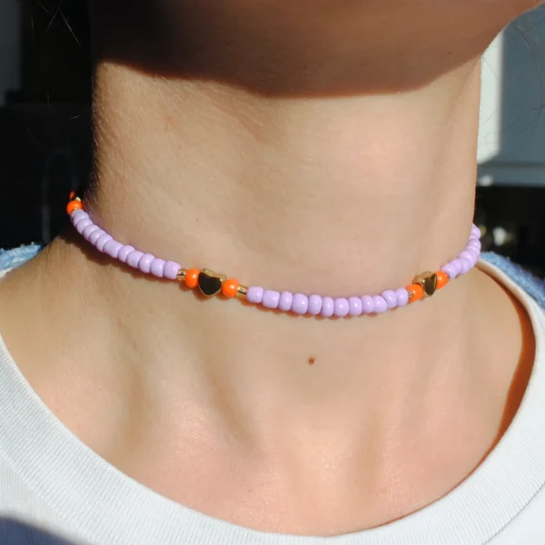 Une personne porte un collier ras-de-cou composé de perles de rocaille violettes, entrecoupées de perles orange vif et de petites breloques en forme de cœur doré. Le collier est ajusté autour du cou, capturant la lumière du jour, ce qui fait ressortir l’éclat des perles et le contraste des couleurs. La personne porte un t-shirt blanc, mettant en valeur les teintes pop et le style délicat du bijou.