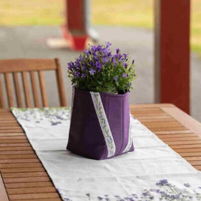 Cache-pot en tissu violet foncé avec galon lavande, posé sur un chemin de table assorti à motif fleuri, sur une table de jardin.