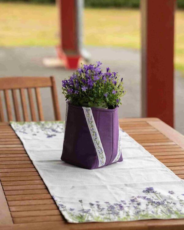 Cache-pot en tissu violet foncé avec galon lavande, posé sur un chemin de table assorti à motif fleuri, sur une table de jardin.
