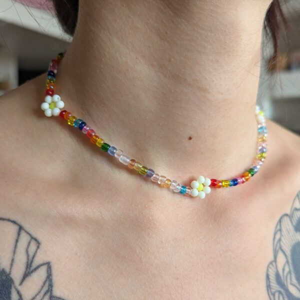 L’image présente un collier ras du cou porté autour d’un cou féminin, avec en arrière-plan une ambiance intérieure douce et floue. Le collier est composé de petites perles colorées translucides et opaques formant un motif multicolore dynamique. Trois petites fleurs blanches en perles sont réparties harmonieusement le long du collier, ajoutant une touche délicate et poétique. Un tatouage floral en noir et gris orne subtilement l’épaule de la personne, renforçant le style bohème de la scène.