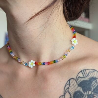 L'image montre un gros plan d'un cou orné d’un collier fait main en perles. Le collier est composé de petites perles multicolores transparentes et opaques, formant un motif arc-en-ciel. Il est ponctué de petites fleurs blanches faites de perles rondes blanches, avec un cœur central jaune ou coloré. Le style du collier est délicat, joyeux, et évoque une esthétique artisanale et estivale. On aperçoit partiellement un tatouage floral noir sur l'épaule de la personne portant le collier.