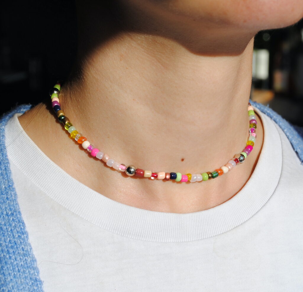 Sur cette image, un ras de cou coloré orne délicatement le cou d’une personne, ajoutant une touche de fantaisie et d’originalité à son style. Le collier est composé d’un mélange harmonieux de perles aux couleurs vives et pastel, incluant du rose, du vert, du jaune, du bleu, du blanc et du rouge. Chaque perle, en perle de rocaille ou plastique comme des boules à facette, est soigneusement agencée pour créer un effet ludique et tendance. Le bijou repose élégamment sur la peau, soulignant la finesse du cou avec son design épuré et ajusté. On y remarque également quelques perles métalliques qui apportent une subtile touche de brillance et de modernité. Ce ras de cou s’inscrit parfaitement dans les tendances bohèmes et Y2K, idéal pour accessoiriser une tenue estivale, décontractée ou même plus sophistiquée en superposition avec d’autres colliers.