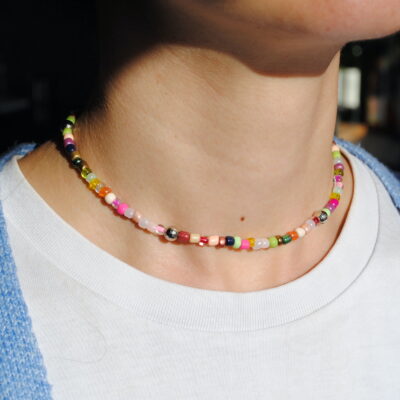 Sur cette image, un ras de cou coloré orne délicatement le cou d’une personne, ajoutant une touche de fantaisie et d’originalité à son style. Le collier est composé d’un mélange harmonieux de perles aux couleurs vives et pastel, incluant du rose, du vert, du jaune, du bleu, du blanc et du rouge. Chaque perle, en perle de rocaille ou plastique comme des boules à facette, est soigneusement agencée pour créer un effet ludique et tendance. Le bijou repose élégamment sur la peau, soulignant la finesse du cou avec son design épuré et ajusté. On y remarque également quelques perles métalliques qui apportent une subtile touche de brillance et de modernité. Ce ras de cou s’inscrit parfaitement dans les tendances bohèmes et Y2K, idéal pour accessoiriser une tenue estivale, décontractée ou même plus sophistiquée en superposition avec d’autres colliers.