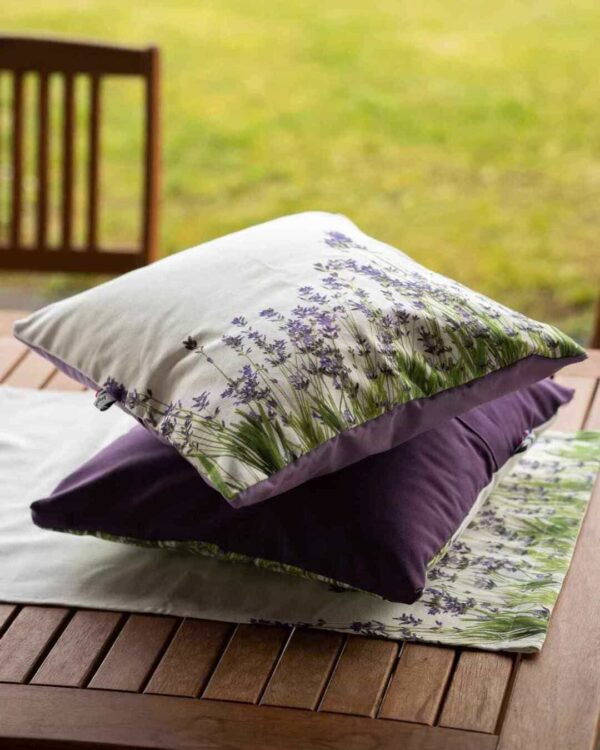 Deux coussins 40 x 40 cm, l’un en tissu imprimé lavande, l’autre avec un côté uni violet, posées sur une table en bois en extérieur.