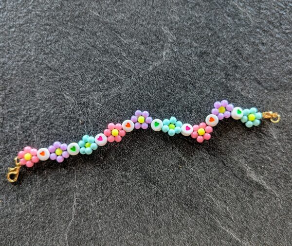 Bracelet posé à plat sur un fond gris foncé. Il est composé de perles en forme de fleurs aux couleurs pastel (rose, violet, bleu, vert) et de perles blanches décorées de petits cœurs colorés (rouge, vert, rose, orange). Le motif alterne entre fleurs et cœurs, formant une ligne ondulée. Les extrémités sont munies de fermoirs dorés.