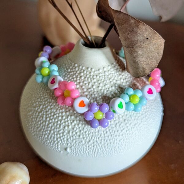 Un bracelet vif et délicat. Il est composé de petites perles arrangées en fleurs multicolores (dans des tons pastel : rose, violet, bleu, vert, jaune), ponctuées de perles blanches décorées de jolis cœurs aux couleurs variées (orange, rose, vert, rouge). Le tout se termine par un fermoir doré accompagné d’un anneau pour l’ajustement.