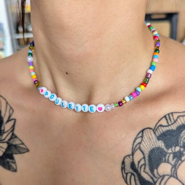 Collier fantaisie fait main porté autour du cou, composé de perles colorées et de lettres formant le mot "POULETTE", encadré par deux cœurs roses. Le collier est associé à un look estival avec un débardeur orange et des tatouages floraux visibles sur la peau.