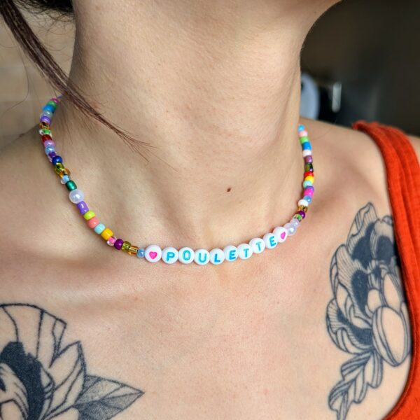 Collier artisanal fait main porté autour du cou, composé de perles colorées et de lettres formant le mot "POULETTE", encadré par deux perles roses en forme de cœur.