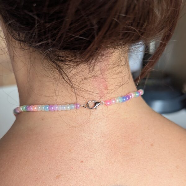 Collier ras de cou porté de dos, sur un cou féminin, composé de perles nacrées multicolores dans des tons pastel (bleu, violet, rose, blanc). Fermoir argenté.