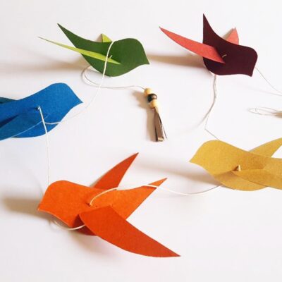 Guirlande décorative faite main composée de cinq oiseaux en papier recyclé de différentes couleurs vives (bleu, vert, rouge, orange et jaune moutarde), suspendus sur un fil blanc avec petites perles en bois et pompon en papier.