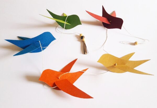 Guirlande décorative faite main composée de cinq oiseaux en papier recyclé de différentes couleurs vives (bleu, vert, rouge, orange et jaune moutarde), suspendus sur un fil blanc avec petites perles en bois et pompon en papier.