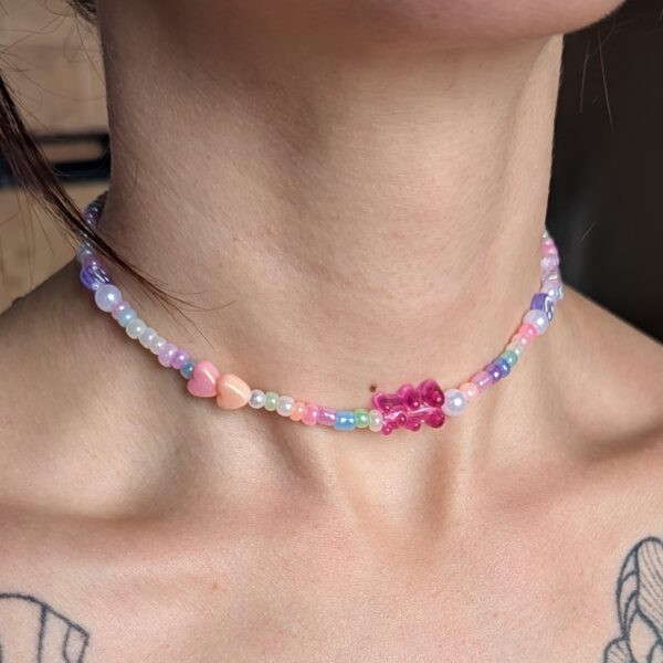 Collier ras de cou composé de perles multicolores pastel, incluant un ourson rose translucide au centre, entouré de perles nacrées, iridescentes et de petits cœurs roses. Le bijou repose délicatement sur la peau, soulignant le cou avec douceur et originalité.