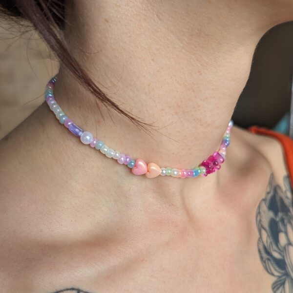 Collier ras de cou fait main, composé de perles pastel irisées dans les tons violets, roses, blancs et bleus. Deux perles en forme de cœur (rose et pêche) sont visibles sur le côté, avec un ourson rose fuchsia au centre. Le collier repose délicatement sur la base du cou.