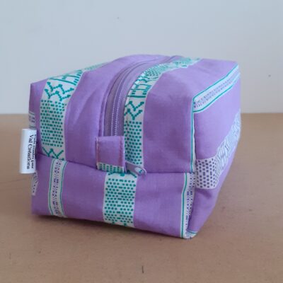 Trousse de toilette zippée, mauve avec de larges bandes blanches et motifs géométrique vert apposées sur celles ci.