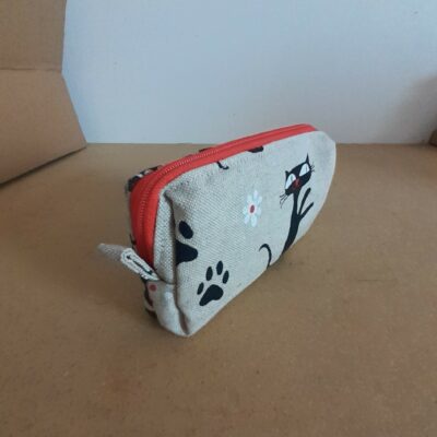 "Une trousse en tissu beige clair avec des motifs de chats noirs, d'empreintes de pattes et d'une petite fleur blanche. La trousse a une fermeture éclair rouge vif et est posée sur une table.