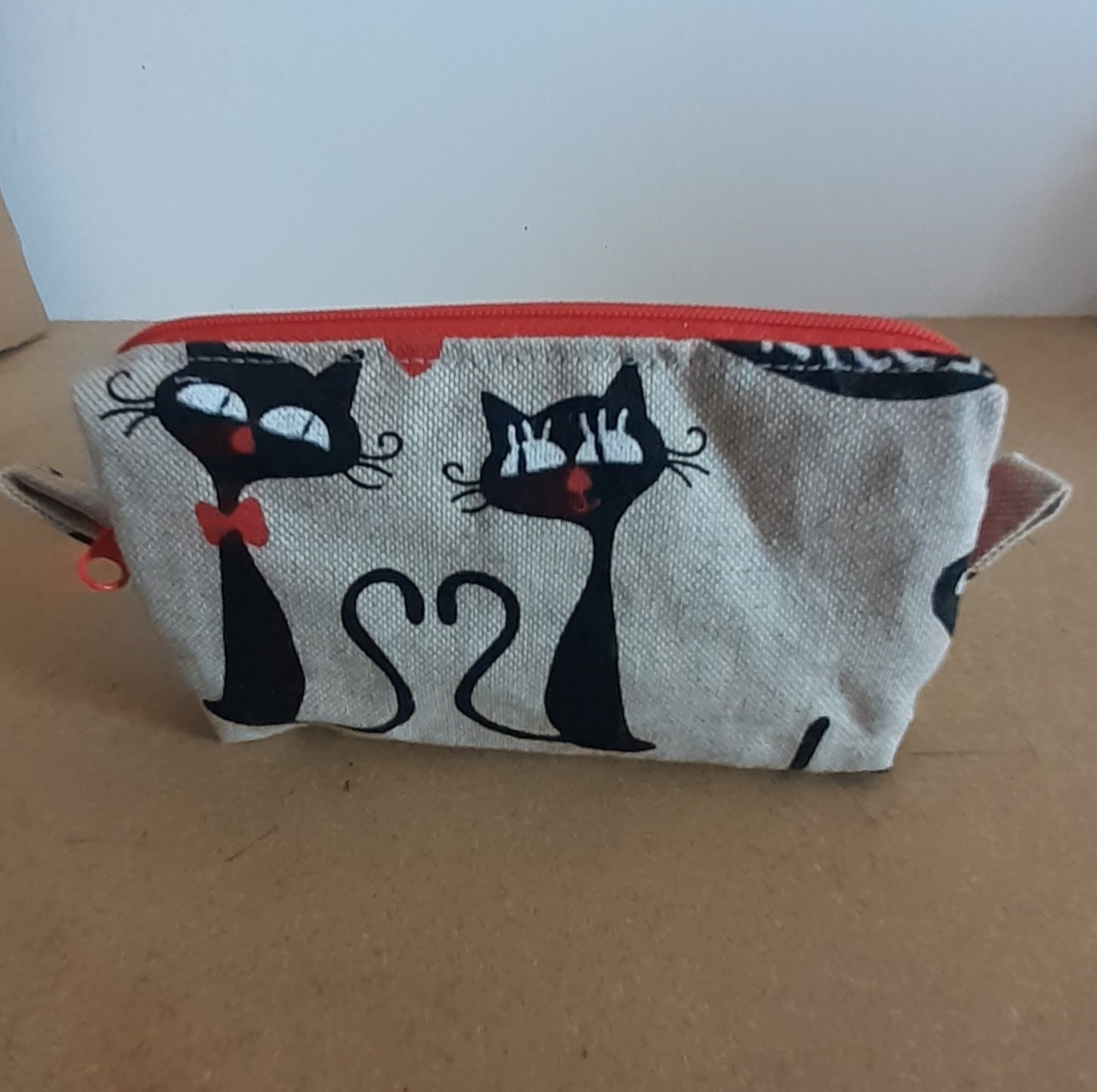"Une trousse en tissu beige clair avec des motifs de chats noirs, d'empreintes de pattes et d'une petite fleur blanche. La trousse a une fermeture éclair rouge vif, posée sur une table vue de face"