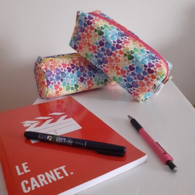 "Deux trousses en tissu coloré à motifs cœurs, posées sur une table blanche à côté d'un carnet rouge et deux stylos."