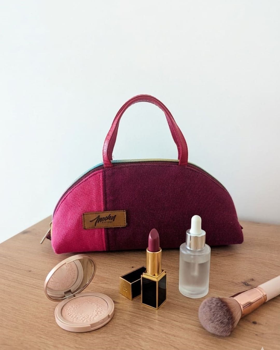 Trousse à maquillage zippée avec anses couleur bordeaux fuchsia avec prodits maquillage posés devant.