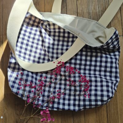 Tote bag upcyclé à carreaux bleu nuit et blanc, posé sur un sol en bois clair, accompagné d’une branche de gypsophile rose.