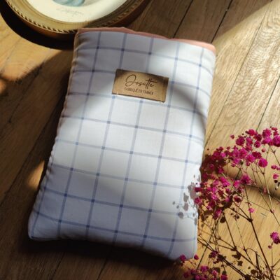 Pochette à livre upcyclée en tissu à carreaux bleu pâle et blanc, posée sur une table en bois, avec un cadre doré et des fleurs roses en décoration.