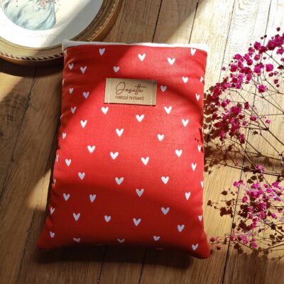 Pochette à livre upcyclée en tissu rouge à petits cœurs blancs, posée sur une table en bois, avec un cadre doré et des fleurs roses en décoration.