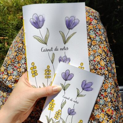 Une main tenant deux carnets A6 et A5 avec une couverture illustrée de fleurs violettes et de boutons d’or peints à l’aquarelle. Le fond est flou, mettant en valeur le carnet.