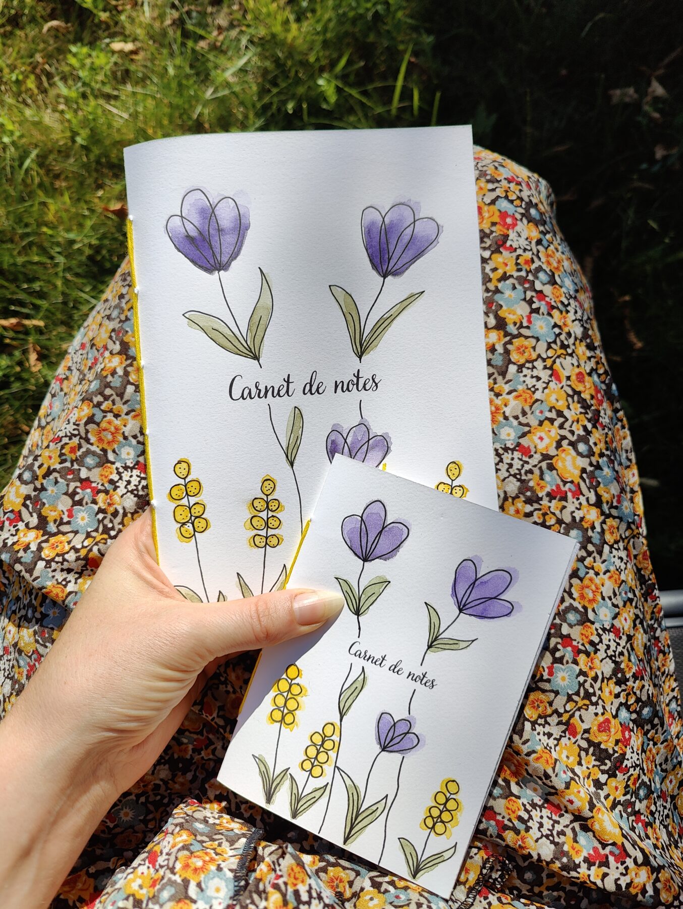 Une main tenant deux carnets A6 et A5 avec une couverture illustrée de fleurs violettes et de boutons d’or peints à l’aquarelle. Le fond est flou, mettant en valeur le carnet.