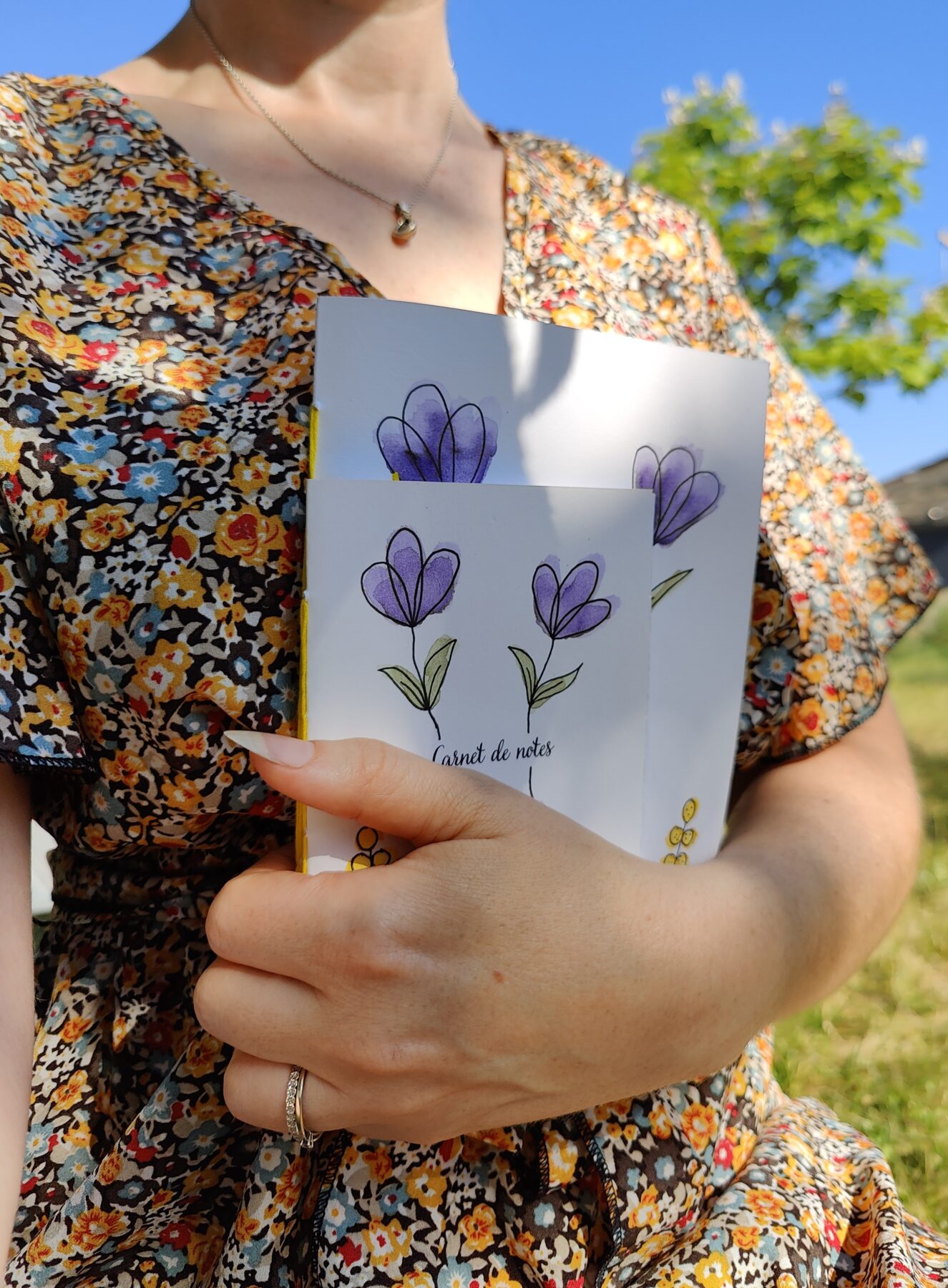 Une main tenant un lot de deux carnets A6 et A5 avec une couverture illustrée de fleurs violettes et de boutons d’or peints à l’aquarelle.