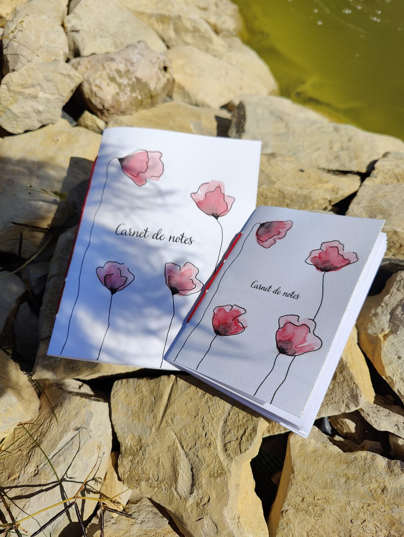 Deux carnets de notes A5 et A6 posés sur des pierres, avec des couvertures illustrées de coquelicots peints à l'aquarelle. Le fond montre un plan d'eau et des pierres naturelles.