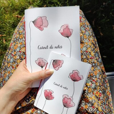 Un carnet de notes A5 et A6 posé sur les genoux d'une personne, avec une couverture illustrée de coquelicots peints à l'aquarelle.