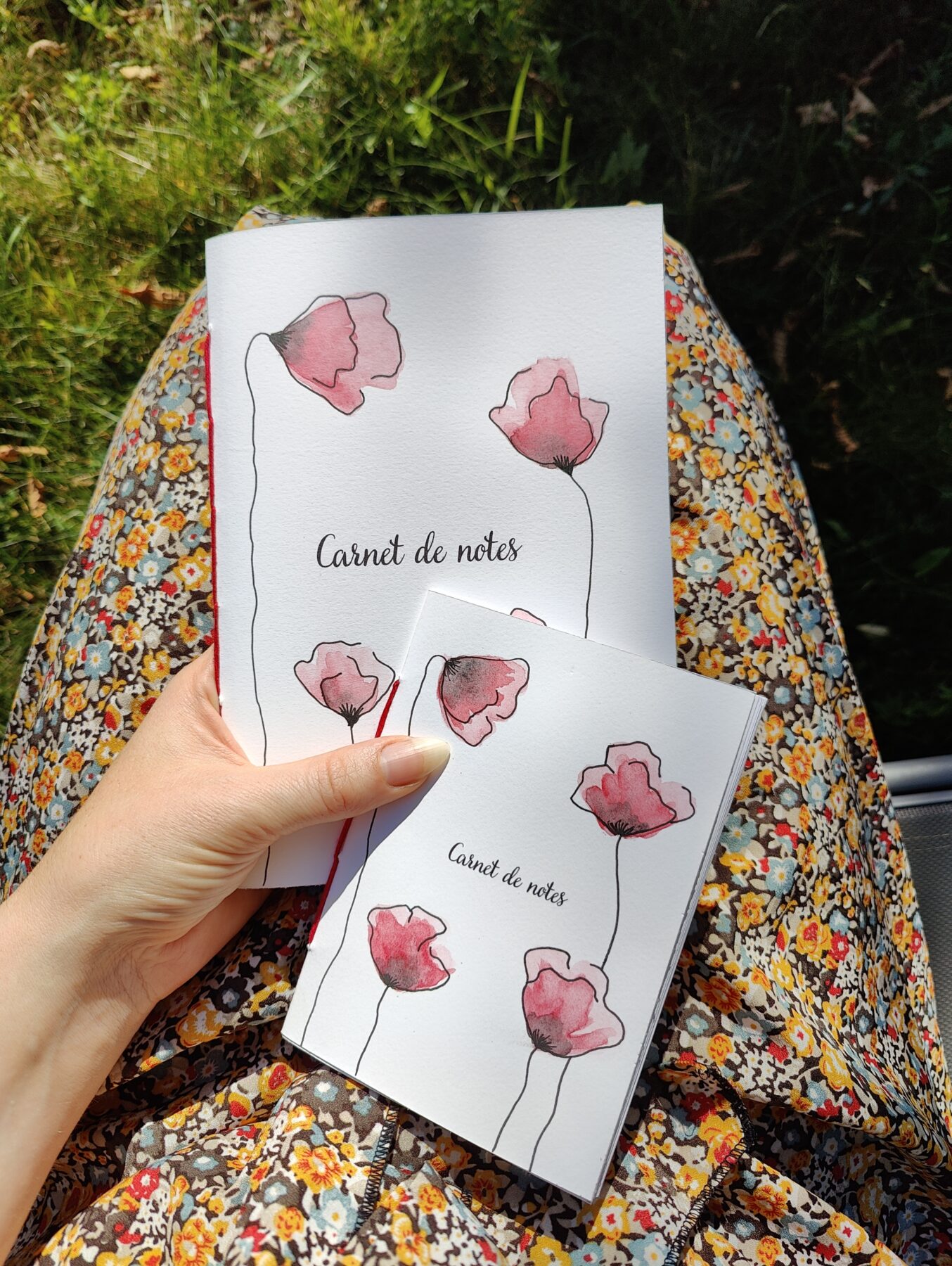 Un carnet de notes A5 et A6 posé sur les genoux d'une personne, avec une couverture illustrée de coquelicots peints à l'aquarelle.