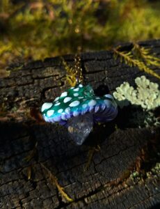 Qu'est-ce que c'est : Un magnifique pendentif artisanal en forme de champignon magique dont la tige est un véritable cristal de quartz brut. Dimensions : Pendentif de 2 × 4 cm, fourni avec une chaîne de 45 cm. Matériaux : Polymère modelé à la main, cristal de quartz naturel et résine époxy de haute qualité. Couleurs : Nuances profondes de vert émeraude, de bleu cosmique et de blanc lumineux. Emballage : Soigneusement emballé dans une jolie boîte cadeau prête à offrir ou à collectionner. Particularités : Le chapeau du champignon est magique : il brille intensément dans le noir et réagit spectaculairement sous l'éclairage de la lumière noire (UV). Styles : Psychédélique, festival, rave, forestier, féerique, fairycore, cottagecore, bohème et mystique.
Laissez-vous enchanter par cette pièce unique inspirée des forêts mystérieuses et des univers oniriques, parfaite pour parfaire votre look de festival ou ajouter une touche de magie à votre quotidien.